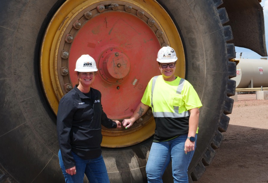 Two women in hardhats and safety vests posing for a photo next to a giant tire
